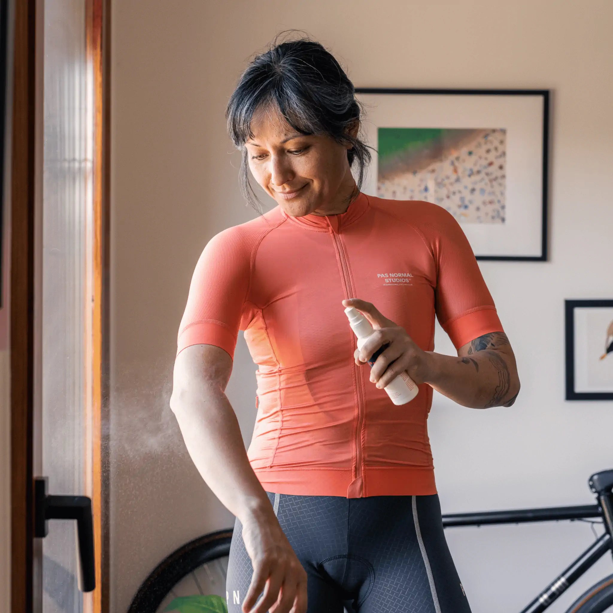 Person wearing an orange cycling jersey applying Pelotan 100ml spray with a bicycle in the background