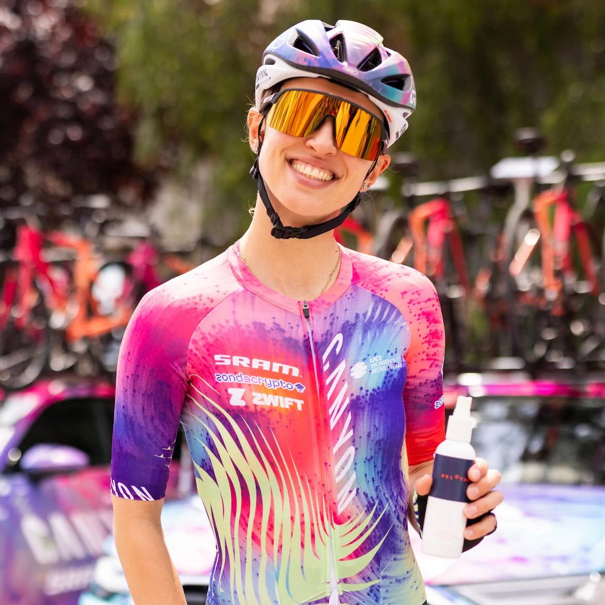 Kasia Niewiadoma from Canyon SRAM Cycling is holding a bottle of Pelotan performance sunscreen beside the team car at the start of a race.