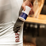 A cyclist is putting a 50ml Roll On bottle of Pelotan Performance Sunscreen into their Rapha cycling jersey back pocket.